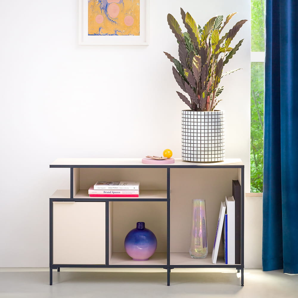 A petite Sand and Midnight Blue Type02 Sideboard with slim legs and mostly open compartments stands decorated with a vase of luscious flowers in front of a white living room wall.