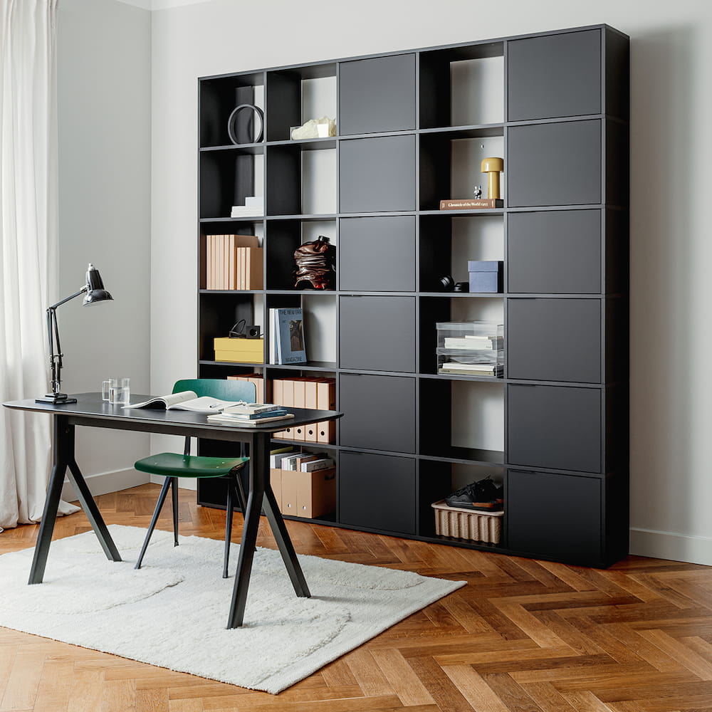 A tall, balanced Type02 Wall Storage in Matte Black stands in a minimalistic office with alternating rows of open and closed storage for displaying and concealing office supplies.